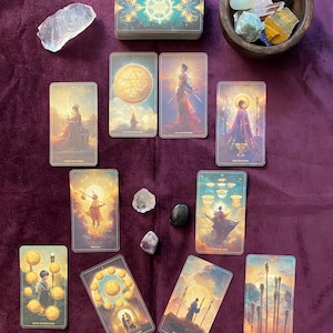 May include: A tarot card spread featuring a deck of cards with celestial and mystical illustrations. The cards are arranged on a purple velvet surface, alongside crystals and a wooden bowl filled with stones. The cards depict various figures and symbols.