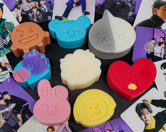 May include: A collection of colorful, handmade soap bars featuring various cartoon characters. The soaps are arranged on a table with several photos of a K-pop group in the background.