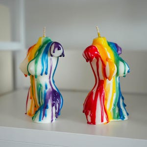 May include: Two white torso-shaped candles featuring a vibrant rainbow-colored wax drip pattern. Each candle has a single wick at the top and displays a multi-colored design with streaks of red, orange, yellow, green, blue, and purple wax.