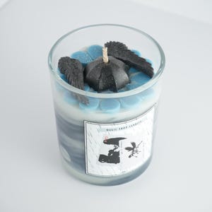 May include: A glass candle with a layered design of blue, white, and gray. The candle is topped with blue wax accents, a black umbrella, and black wing accents. The label reads "MAGIC SHOP CANDLES" with a graphic.