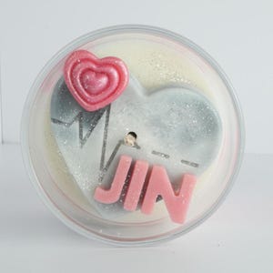 May include: A candle with a heart-shaped design, featuring a pink heart and the name "JIN" in pink letters. The candle has a heartbeat line and is encased in a clear container. The candle has a glittery appearance.