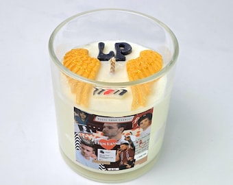 May include: A white candle in a clear glass jar with a collage of photos on the label. The candle has two yellow wings and the letters "LP" on top. The label says "Magic Shop Candles".