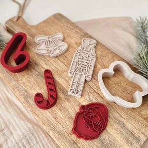 May include: A collection of six holiday-themed cookie cutters in red and off-white. The set includes a candy cane, a nutcracker, a pair of bells, a Santa Claus face, and two abstract shapes, all arranged on a wooden cutting board surface.