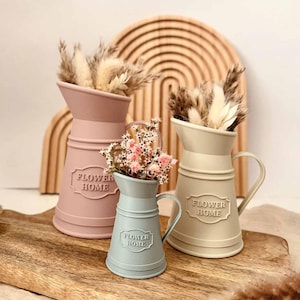May include: Three pastel-colored metal pitchers with dried flowers. The pitchers are in pink, light green, and beige. Each pitcher has a handle and the words "FLOWER HOME" embossed on the front. They are arranged on a wooden cutting board.