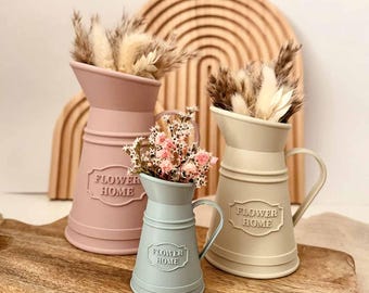 Puede incluir: Tres jarrones de metal con forma de jarra en rosa empolvado, azul claro y crema. Cada jarrón tiene un asa, un borde acampanado y una placa rectangular en relieve en la parte frontal con el texto FLOWER HOME. Están llenos de hierbas y flores secas.
