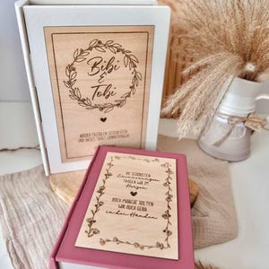 Könnte beinhalten: Eine weiße buchförmige Box mit einer hellen Holzplatte, die einen Kranz und den Text Bibi & Tobi zeigt, sowie eine kleinere rosafarbene buchförmige Box mit einer hellen Holzplatte, floralem Rand und deutschem Text über Erinnerungen und Herzen.
