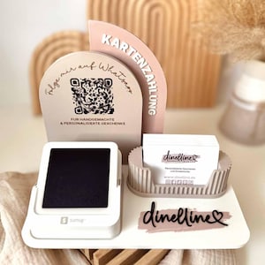 May include: A white point-of-sale system with a black screen, a business card holder, and decorative signs. The signs include a QR code and the text "Follow me on Whatsapp" and "Card Payment". The business card holder has the text "dinelline" on the cards.