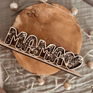 May include: Wooden sign with the word "MAMA" in a decorative font. The letters are cut out and have a floral design. The sign is on a wooden base.