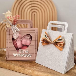 Puede incluir: Una caja de regalo rosa polvoriento con forma de bolso y un recorte en forma de corazón, que contiene objetos rosas y el texto DU BIST MEIN MENSCH, junto a una caja de regalo blanca moteada con forma de bolso y un lazo hecho de papel moneda.