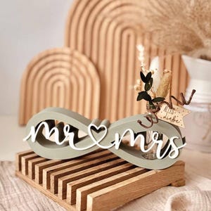 May include: A sage green infinity-shaped decorative object featuring the white cursive text "mr & mrs" with a heart symbol in the centre. A small glass vase with dried flowers and a wooden tag reading "Alles Liebe" is tucked into the infinity loop.