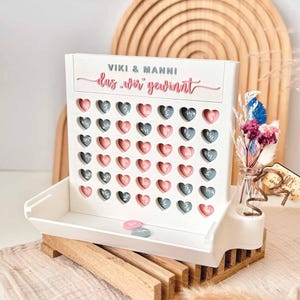 May include: A white wooden game board with a grid of heart-shaped cutouts, featuring the text VIKI & MANNI and das wir gewinnt in pink and grey. The board includes pink and grey heart tokens, a small glass vase with dried flowers, and a wooden base.