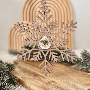 Puede incluir: Un adorno de copo de nieve de madera cortado con láser que presenta un diseño simétrico de seis puntas con detalles intrincados. Un cristal transparente, facetado y en forma de lágrima está suspendido en el centro por una pequeña anilla metálica.