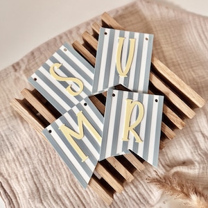 May include: Four rectangular pennants with a light blue and white vertical striped pattern. Each pennant features a single, raised, pale yellow letter in the center, spelling out the letters S, U, M, and R, with two small holes at the top.