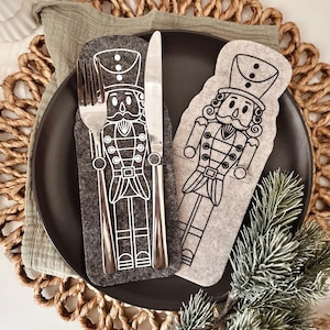 May include: A black plate with a fork and knife set on a gray felt nutcracker-themed utensil holder. The holder has a black outline of a nutcracker soldier. The plate is on a woven placemat with a green cloth napkin.