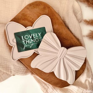 Könnte beinhalten: Ein hellrosa schleifenförmiger Geschenkkartenhalter und eine passende schleifenförmige Dekoration. Der Geschenkkartenhalter hat eine grüne Karte mit dem Text "LOVELY THINGS for you". Die Artikel befinden sich auf einem Holztablett.