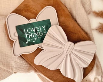 Puede incluir: Un tarjetero en forma de lazo rosa claro y una decoración a juego en forma de lazo. El tarjetero tiene una tarjeta verde con el texto "LOVELY THINGS for you". Los artículos están sobre una bandeja de madera.