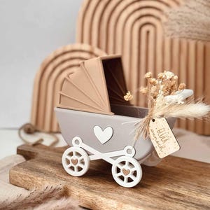 Può includere: Una carrozzina decorativa in legno grigio e bianco con un dettaglio a cuore e una capote beige. La carrozzina ha ruote bianche con cuori intagliati ed è adornata con fiori secchi e un'etichetta di legno con la scritta "Glückwunsch".