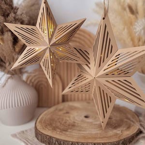 Könnte beinhalten: Zwei hängende sternförmige Ornamente aus hellbraunem Material mit komplizierten geometrischen Ausschnitten. Jeder Stern hat ein hohles Zentrum, das durch interne Lichter ein warmes Leuchten abgibt und an dünnen Schnüren hängt.