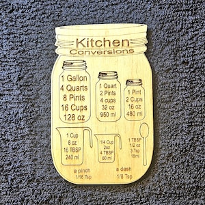 May include: A wooden kitchen conversion chart shaped like a mason jar. The chart includes measurements for gallons, quarts, pints, cups, ounces, and milliliters. It also includes measurements for cups, tablespoons, teaspoons, and ounces. The chart is a helpful tool for cooks and bakers.
