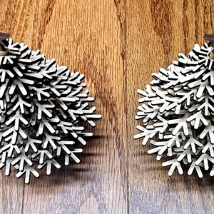Può includere: Due decorazioni natalizie in legno a forma di albero. Ogni albero è bianco con bordi neri e una piccola stella marrone in cima. Le decorazioni sono su una superficie di legno.