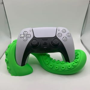 May include: A white and black PlayStation 5 controller is cradled by a bright green octopus tentacle-shaped stand. The stand has suction cup details and is designed to hold the controller.