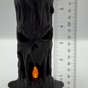 May include: A black, cylindrical, sculpted candle holder featuring a menacing, screaming face with deep-set eyes and an open mouth. A small, orange, flame-shaped LED light is visible inside the mouth. A ruler shows a height of 5.5 inches.
