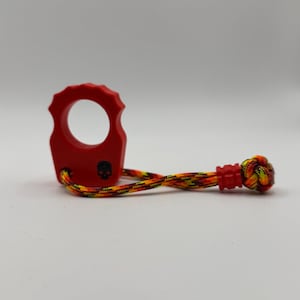 May include: A red finger ring tool with a scalloped outer edge and a small black skull icon printed on the front. A multicolored paracord lanyard in shades of red, yellow, and black is attached to the base, featuring a knotted end and a red bead.
