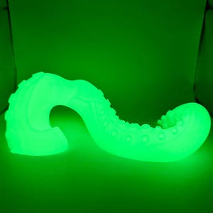May include: A glowing green, curved tentacle-shaped object with textured details. The object has a series of rounded bumps along its length and a textured, ridged surface. The overall shape is reminiscent of an octopus tentacle.