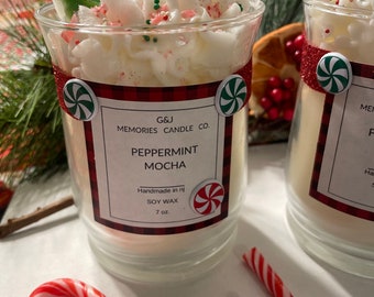 May include: Two peppermint mocha scented soy wax candles in glass jars with white whipped cream and red and green sprinkles on top. The candles are decorated with a candy cane and a green holly sprig. The jars have red and black plaid labels with the text "G&J Memories Candle Co. Peppermint Mocha Handmade in NJ Soy Wax 7 oz."