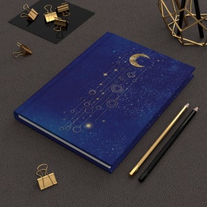 Puede incluir: Un cuaderno azul con un diseño celestial dorado que presenta una luna creciente, estrellas y planetas. El cuaderno está sobre una superficie gris con dos lápices y clips de papel dorados.