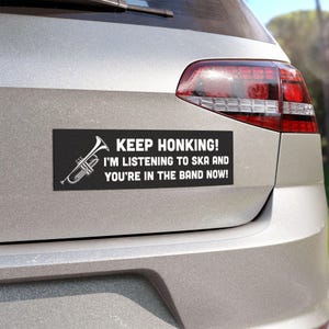 以下が含まれることがあります： 「Keep Honking! I'm listening to ska and you're in the band now!」と書かれた白いテキストが書かれた黒いバンパーステッカー。ステッカーの左側には白いトランペットのグラフィックがあります。