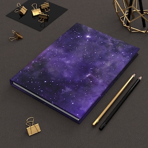 Op de afbeelding: Een paars notitieboek met een galaxy-printontwerp. Het notitieboek ligt op een donkergrijs oppervlak met gouden paperclips en potloden.