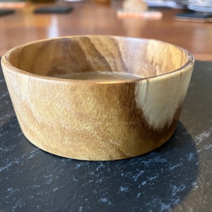 Puede incluir: Un pequeño cuenco de madera redondo de color marrón claro con un patrón de veta de madera natural. El cuenco está hecho de madera maciza y tiene un acabado liso y pulido.