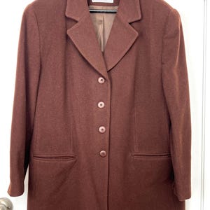 May include: A solid dark brown blazer featuring a notched lapel, four matching buttons down the front, and two horizontal welt pockets. The interior label reads Holt Renfrew Classics. The garment is displayed on a black hanger against a white door.