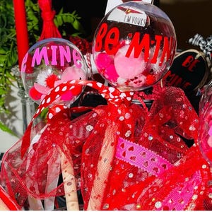 Puede incluir: Dos adornos de vidrio transparente con pompones rojos y rosas y purpurina en el interior. Los adornos están decorados con letras rojas de vinilo que dicen "Be Mine" y "I'm Yours". Los adornos están unidos a palos y envueltos en tul rojo y blanco.