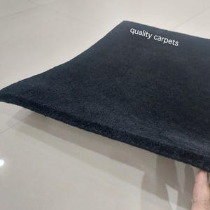 May include: A thick, solid black rectangular carpet with a dense, textured pile surface. The text "quality carpets" is printed in white on the upper portion of the carpet. The material appears firm and uniform in color throughout the entire piece.