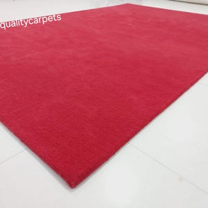 May include: A rectangular area rug in a solid, vibrant red color with a short, dense pile texture. The text qualitycarpets is printed in white in the upper left corner of the rug. The rug is positioned at an angle on a plain white surface.