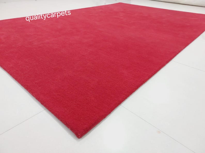 May include: A rectangular area rug in a solid, vibrant red color with a short, dense pile texture. The text qualitycarpets is printed in white in the upper left corner of the rug. The rug is positioned at an angle on a plain white surface.