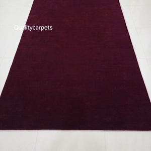 May include: A rectangular area rug in a deep, solid burgundy color with a smooth, uniform texture. The text "Qualitycarpets" is printed in white on the left side of the rug. The rug is positioned vertically, displaying a consistent dark red hue.