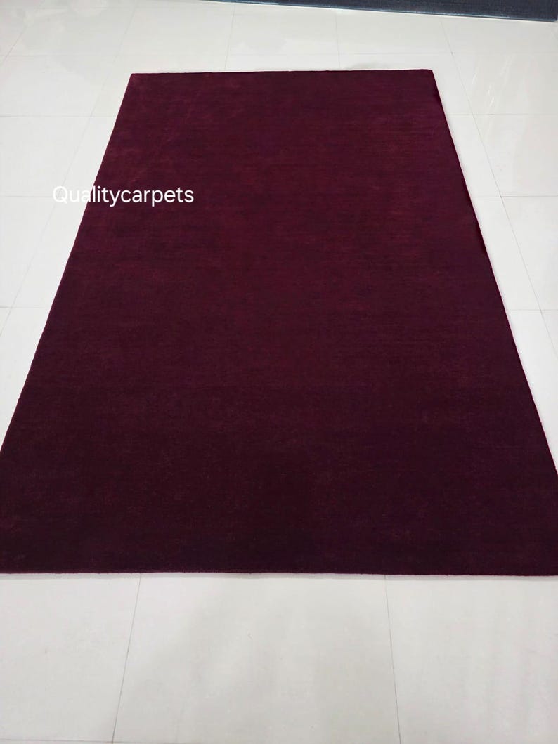 May include: A rectangular area rug in a deep, solid burgundy color with a smooth, uniform texture. The text "Qualitycarpets" is printed in white on the left side of the rug. The rug is positioned vertically, displaying a consistent dark red hue.