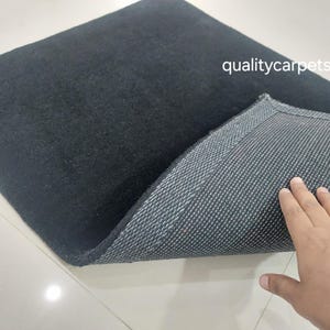 May include: A square black rug with a soft, solid-colored top surface and a textured, dark gray non-slip backing. The text qualitycarpets is printed in white on the upper right side of the rug, which is partially folded to reveal the underside.