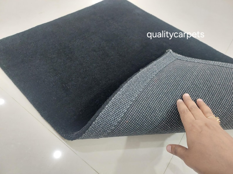May include: A square black rug with a soft, solid-colored top surface and a textured, dark gray non-slip backing. The text qualitycarpets is printed in white on the upper right side of the rug, which is partially folded to reveal the underside.