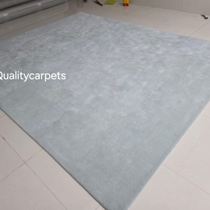May include: A large, square, light blue area rug with a soft, uniform texture. The surface of the rug features a subtle, mottled appearance in varying shades of pale blue. The text "Qualitycarpets" is printed in white on the left side of the rug.