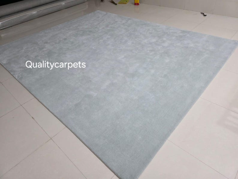 May include: A large, square, light blue area rug with a soft, uniform texture. The surface of the rug features a subtle, mottled appearance in varying shades of pale blue. The text "Qualitycarpets" is printed in white on the left side of the rug.