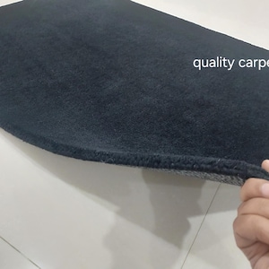 May include: A rectangular black carpet with a thick, plush pile texture. The text "quality carpets" is printed in white on the surface. One corner of the carpet is lifted to reveal a gray, textured backing material underneath the black fibers.