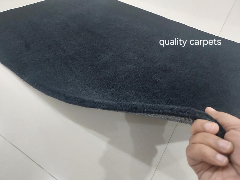 May include: A rectangular black carpet with a thick, plush pile texture. The text "quality carpets" is printed in white on the surface. One corner of the carpet is lifted to reveal a gray, textured backing material underneath the black fibers.