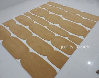 May include: A beige and white area rug with a geometric pattern of rounded ovals. The rug has a modern design and is made of soft, plush fibers. "quality carpets" is printed in the bottom right corner of the rug.