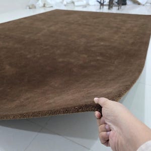 May include: A rectangular area rug in a solid dark brown color. The rug features a dense, short-pile texture across its entire surface. One edge of the rug is lifted, revealing the thickness and the consistent brown material of the backing.