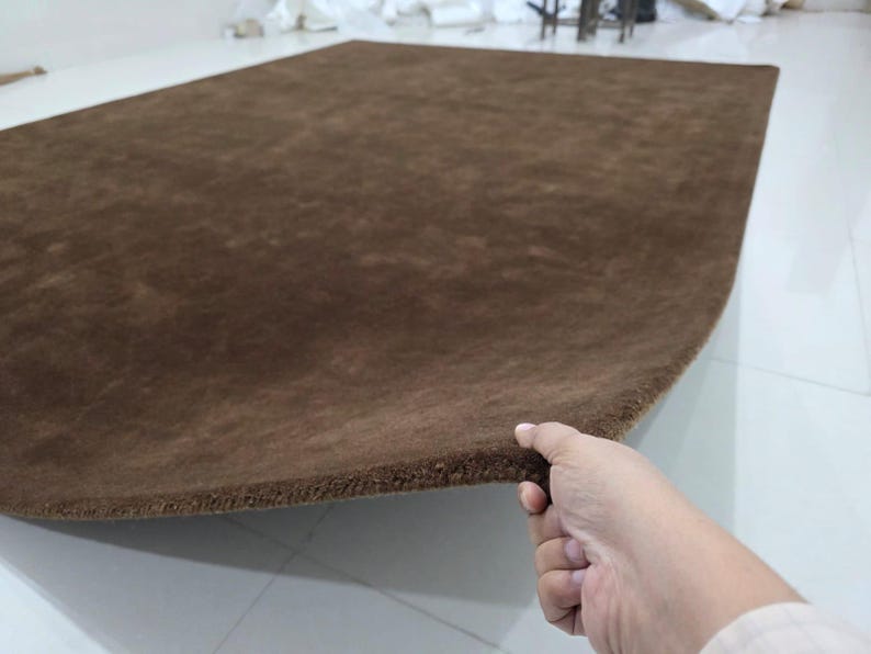 May include: A rectangular area rug in a solid dark brown color. The rug features a dense, short-pile texture across its entire surface. One edge of the rug is lifted, revealing the thickness and the consistent brown material of the backing.