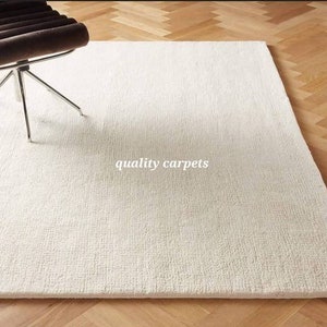 May include: A rectangular off-white area rug with a textured, woven surface. The rug features the text quality carpets printed in white in the center. A portion of a dark brown chair with a chrome metal base is visible in the upper left corner.